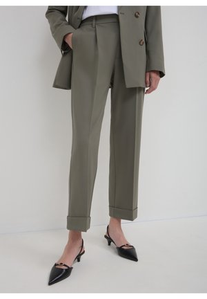 Femme portant un pantalon vert olive sur mesure avec des ourlets retournés et des chaussures slingback noires pointues à talon bas, debout devant un fond uni.