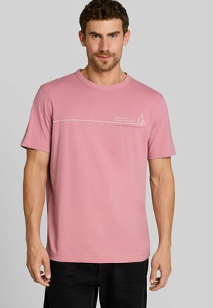 Mann trägt ein hellrosa T-Shirt mit kurzen Ärmeln, weißem Text und einer kleinen Segelboot-Grafik auf der oberen linken Brust.