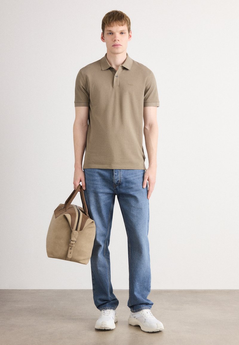 Jeune homme portant un polo marron, un jean bleu, des baskets blanches, tenant un sac de sport beige avec des anses marron, se tenant sur un sol gris.