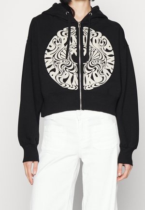 Personne portant un sweat à capuche noir court avec fermeture éclair et un design circulaire abstrait blanc, ainsi qu'un pantalon blanc, debout devant un fond uni.