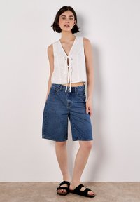 Weiße, ösenmusterige, ärmellose Bluse mit V-Ausschnitt und Schleifenfront, kombiniert mit blauen Jeans-Shorts und schwarzen Rutsch-Sandalen.