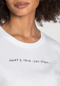 Camiseta de algodón blanca con un cuello redondo y un diseño de texto impreso en negro que dice "¿CUÁL ES TU HISTORIA DE AMOR?"