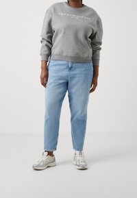 Harmaa hupullinen paita, jossa on valkoinen logo, vaaleansiniset farkut ja hopeiset lenkkarit. Ominaisuuksina rento istuvuus ja taitetut hihansuut. Yksivärinen tausta.