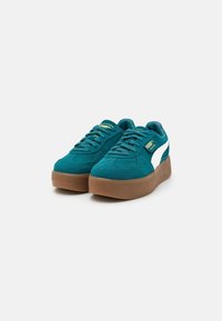 Teal suede sneakers with a white logo, featuring round laces and a light brown rubber platform sole. Accents include gold branding.