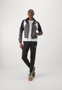 Giacca sportiva grigia e bianca con chiusura a zip, abbinata a pantaloni neri e scarpe da trail multicolori. Tessuto liscio e leggero.