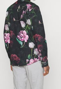 Uomo che indossa una camicia nera a maniche lunghe con un grande motivo floreale in rosa, bianco e rosso, abbinata a pantaloni grigi chiari, visto da dietro.