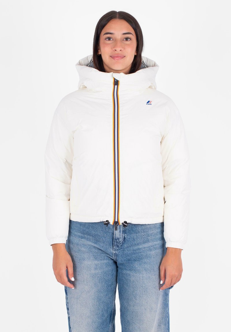 Witte pufferjack met een capuchon, voorzien van een rits aan de voorkant met multikleuren strepen en een trekkoord aan de zoom. Model draagt blauwe spijkerbroek.