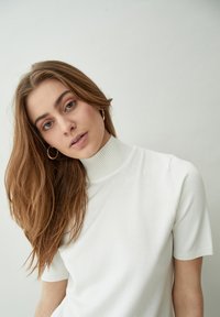 Top de punto blanco de manga corta con cuello acanalado alto. La textura suave y el diseño ajustado realzan la simplicidad y elegancia de la prenda.
