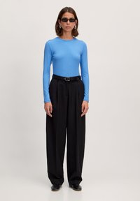 Blauwe top met lange mouwen en ribbelstructuur, gecombineerd met zwarte hoge taillebroek. De look bevat een zwarte riem en zwarte zonnebril.