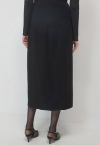 Jupe mi-longue noire avec une texture lisse, un design ajusté et une poche à l'arrière. Portée avec des chaussures à talons noires et des bas transparents.