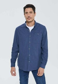 Chemise bleu à boutons dans un tissu doux avec une poche, dotée de boutons en argent et d'un col. Portée avec un t-shirt blanc et un jean.