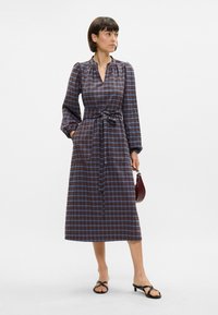 Langermet rutete kjole i mørkeblått og brunt, med belte i midjen, lommer og kragehals, kombinert med svarte sandaler.