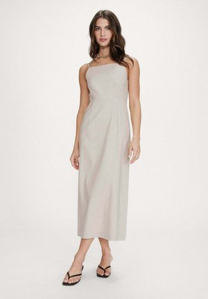 Femme debout portant une robe midi beige sans manches, des sandales noires et un collier chaîne en or, sur un fond clair uni.