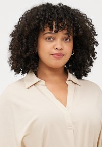 A woman with curly hair and hoop earrings gazes directly at the camera, wearing a light beige collared shirt against a neutral background.