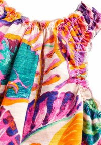 Barevná látka s květinovým vzorem zahrnujícím zelené, růžové, fialové a oranžové odstíny. Detail elastického volánkového výstřihu. Měkká textilie.