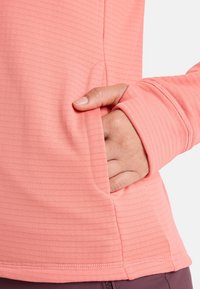 Lightweight, ribbed long-sleeve top in coral, featuring a side pocket and thumbholes in the cuffs, designed for comfort and ease of movement.