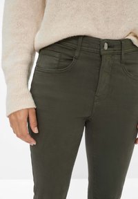 Olivgrüne Slim-Fit-Hosen mit einem Knopfverschluss und dezenten Stickdetails, kombiniert mit einem leichten, strukturierten beigen Pullover.