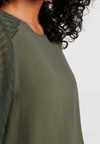 Donna che indossa un top verde oliva con maniche trasparenti a fantasia, capelli ricci scuri in vista, su uno sfondo chiaro e semplice.