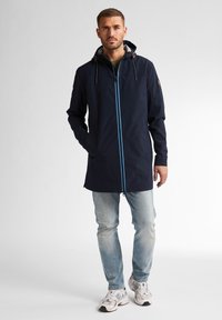 Granatowa wodoodporna kurtka z niebieskim zamkiem, regulowanym kapturem i dwoma przednimi kieszeniami, noszona na jasnoniebieskich dżinsach i białych butach sportowych.