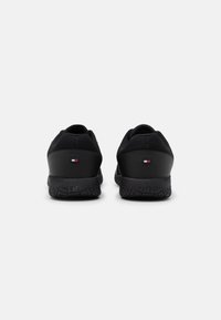 Paire de baskets basses noires avec semelles texturées et petit logo rectangulaire rouge, blanc et bleu sur chaque talon, vues de derrière.
