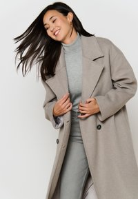 Beige wollen overjas met grote revers en twee knopen. Draagt over een lichtgrijze coltrui en grijze jeans, met een zachte textuur en een op maat gemaakte pasvorm.