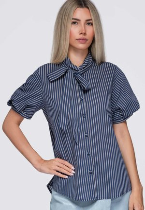 Junge Frau trägt eine marineblaue und weiß gestreifte Bluse mit kurzen Puffärmeln und einer gebundenen Schleife am Hals.