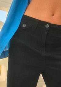 Črne hlače z gladko teksturo, ki imajo na pasu dva siva gumbna zapirala, delno pokrita z modro srajco.