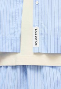 Vêtement rayé verticalement bleu clair et blanc avec des boutons blancs, montrant une étiquette en tissu blanc avec « ROUGE EDITION » imprimé en noir.