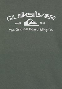 Logo Quiksilver con icona di onda e montagna, testo "Dal 1969, The Original Boardriding Co." su sfondo di tessuto verde scuro.