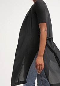 Person trägt eine ärmellose schwarze, offene lange Weste über einem schwarzen T-Shirt mit kurzen Ärmeln und hellblauen Jeans vor einem einfarbigen Hintergrund.