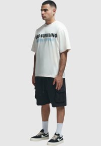 T-shirt oversize blanc avec un texte noir et bleu, associé à un short cargo noir et des baskets noires. Le mannequin se tient de profil gauche.