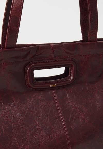 Burgundy leather tote bag with a textured finish, featuring a rectangular cut-out handle and gold "maje" logo. Detailed stitching visible.