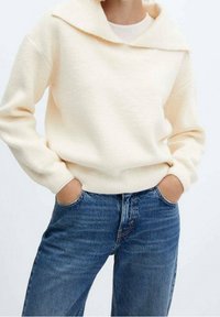 Pull en laine couleur crème avec un large col, des manches longues et des poignets côtelés, associé à un jean bleu taille haute. Design simple.