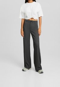 Žena v šedých širokých kalhotách, bílém zkráceném tričku s nařasením a bílých teniskách, stojící před jednobarevným šedým pozadím.