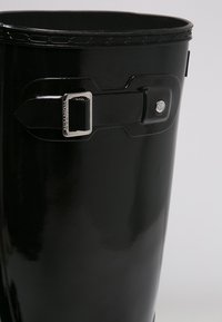 Stivale in gomma nera con finitura lucida, cinturino regolabile con fibbia in metallo e bordo superiore testurizzato. Profilo sottile con design arrotondato.