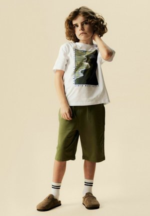 Dziecko z kręconymi włosami, ubrane w białą koszulkę z motywem surfingowym, oliwkowo-zielone szorty, białe skarpetki w paski i brązowe wsuwane buty, stojące z ręką na szyi.