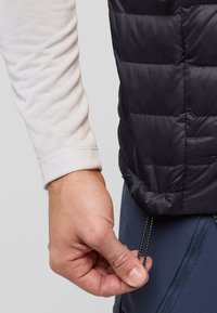 Schwarze Pufferweste mit gestepptem Design und Kordelzug. Helle Langarmbluse darunter. Hand, die den Saum der Weste anpasst.