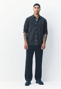 Camicia con bottoni davanti in motivo scuro a quadri, abbinata a pantaloni scuri e scarpe nere. Il tessuto presenta una texture; la camicia ha le maniche arrotolate.