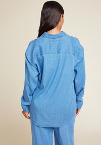 Lys blå denimskjorte med lange ermer, rund halslinje og buet kant. Har synlige sømmer og en avslappet passform.