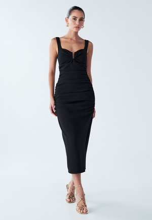Robe noire ajustée avec un décolleté en cœur, des détails froncés et une petite ouverture à l'avant. Portée avec des sandales dorées à lanières.