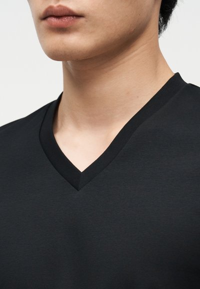 Εγγύς εικόνα του κάτω μέρους του προσώπου και του λαιμού ατόμου που φοράει απλό μαύρο T-shirt με V λαιμόκοψη, πάνω σε ανοιχτόχρωμο φόντο.