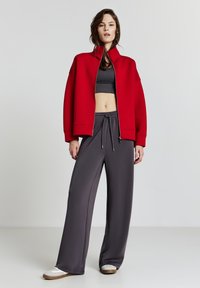 Donna che indossa una giacca rossa zip-up sopra un top corto nero, pantaloni grigio scuro larghi con cordino e sneakers bianche su uno sfondo neutro.