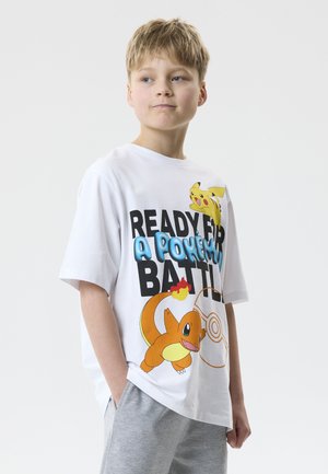 Băiat purtând un tricou alb Pokémon cu grafice Pikachu și Charmander, combinat cu pantaloni gri deschis, stând cu mâinile în buzunare.