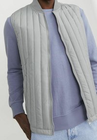 Grå quiltad väst med ribbat krage, dragkedja fram och två sidofickor, bärs över en ljuslila långärmad skjorta.