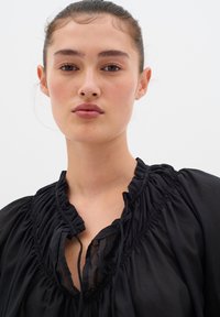 Blusa nera con tessuto arricciato, scollatura a volant e apertura a occhiello. Texture liscia su uno sfondo bianco semplice.