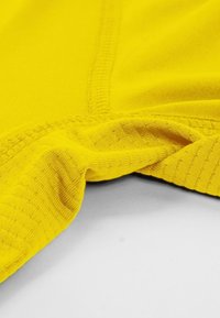 Tela amarilla con una textura suave, que presenta un borde doblado y costuras visibles. El material parece ligero y elástico.