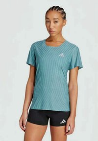 Kvinne med flette som har på seg en blå mønstrete treningsskjorte og svarte shortser med en hvit sportslogo, stående mot en ensfarget bakgrunn.