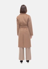 Cappotto in misto lana marrone con vita aderente, collo a punta e maniche lunghe, abbinato a pantaloni beige a gamba larga e tacchi neri.