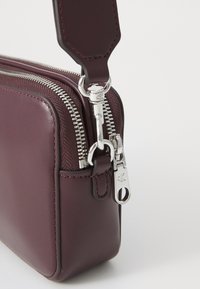 Bordeaux leren tas met zilverkleurige hardware, dubbele ritsluiting en een afneembare schouderriem. Glad textuur met gestikte accenten.
