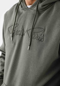 Hoodie vert foncé en tissu doux. Présente un logo sur le devant en fil assorti, une capuche ajustable avec cordons de serrage et embouts en métal.